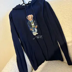 Säljer en snygg mörkblå hoodie från Ralph Lauren med en cool björn på framsidan. Perfekt för en avslappnad stil. Den har långa ärmar och en klassisk huva med dragsko. Passar perfekt till både jeans och joggers. Skön och stilren för alla tillfällen!