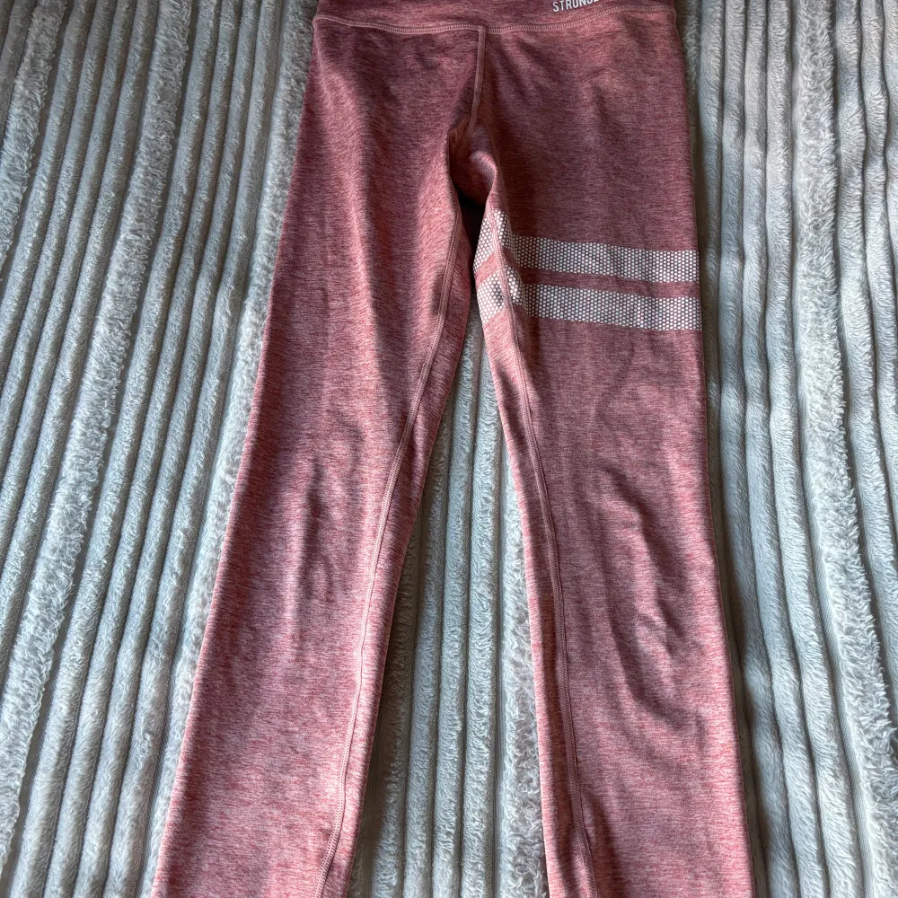 Säljer ett par snygga rosa leggings från Stronger. De är perfekta för träning med sin bekväma passform och stretchiga material. Leggingsen har vita ränder på ena benet som en cool detalj. Perfekta för gymmet eller en löprunda!. Jeans & Byxor.