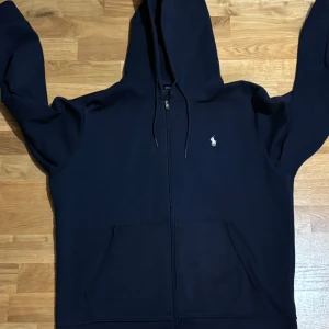 Mörkblå hoodie från Polo Ralph Lauren - Säljer en snygg mörkblå hoodie från Polo Ralph Lauren i storlek L. Den har en dragkedja framtill och en klassisk logga på bröstet. Perfekt för höst och vinter, med en skön passform och praktiska fickor. Huvan har dragsko för justering. Perfekt för en casual look! 💙