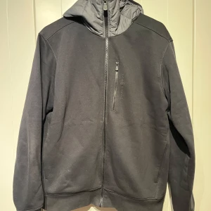 Svart hybridjacka från Sail Racing - Säljer en svart hybridjacka från Sail Racing i storlek XL som passar L. Den har en dragkedja framtill och en huva i dun. Perfekt för höst och vår men kan även användas som hoodie. Jackan är i mycket bra skick. Nypris 1800:-