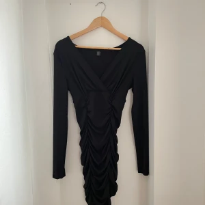 Svart långärmad klänning - Snygg svart klänning med lång ärm och omlott-design. Den har en åtsittande passform och rynkade detaljer framtill som ger en elegant look. Perfekt för festliga tillfällen eller en utekväll. Materialet känns mjukt och stretchigt.