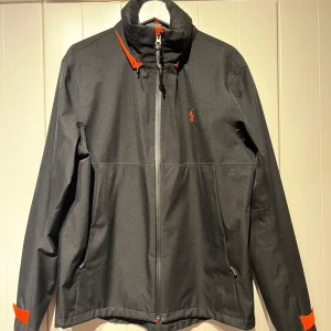 Svart vindjacka från Ralph Lauren - Säljer en snygg samplejacka från Raph Lauren. Jackan är svart med orangea detaljer. Jackan är i mycket bra skick, nypris 2500:-