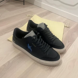 Svarta sneakers från Axel Arigato - Säljer ett par snygga svarta sneakers från Axel Arigato. De har en cool blå fågelbrodering på sidan och är i mycket bra skick. Skorna har en vit sula och klassisk snörning. Perfekta för både vardag och fest!