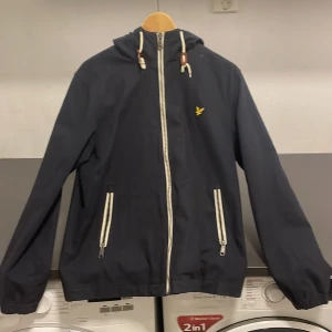 Windbreaker från Lyle & Scott - Säljer en snygg jacka från Lyle & Scott i storlek S. Jackan har en dragkedja framtill och praktiska fickor med dragkedjor. Den har en huva med justerbara snören och är perfekt för höst och vår. Märkets logga syns på bröstet. Perfekt för en stilren look!