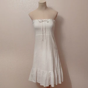 Vit axelbandslös klänning - Säljer en superfin vit axelbandslös klänning med smockdetaljer upptill och en söt rosett framtill. Klänningen har en volangkant längst ner som ger en romantisk touch. Perfekt för sommarens alla fester eller en dag på stranden! Inha flävka eller hål.