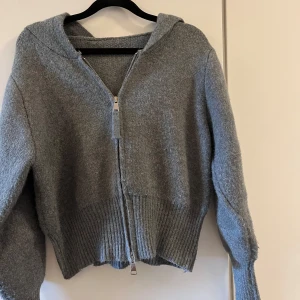Stickad kofta - Stickad kofta med en luva i en croppad modell, den har två dragkedjor. Bra skick, endast lite nopprig på vissa ställen men inget man tänker på. Står ingen storlek men skulle säga S