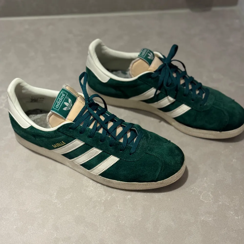 Säljer ett par snygga gröna Adidas Gazelle sneakers i mocka. De har de klassiska tre vita ränderna på sidan och en vit sula. Skorna är i bra skick. . Skor.