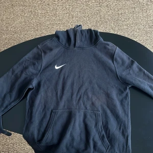 Svart hoodie från Nike - Säljer en svart hoodie från Nike med klassisk design och vit logga på bröstet. Tröjan har en stor ficka framtill och är tillverkad i mjukt material för extra komfort. Perfekt för en avslappnad stil.