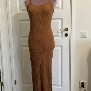 Brun ribbad långklänning -  brun ribbad långklänning med smala axelband och en liten slits på ena sidan av klänning. Tajt passform