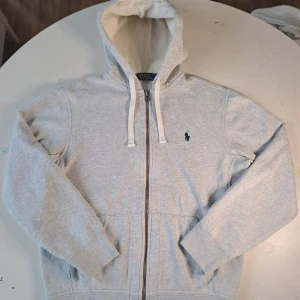 Grå hoodie från Polo Ralph Lauren - Säljer en stilren grå hoodie från Polo Ralph Lauren med dragkedja och klassisk logga på bröstet. Tröjan har en justerbar huva med vita snören och långa ärmar. Perfekt för en avslappnad look. Tröjan är I nyskick 
