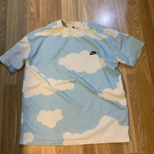 Blå och vit t-shirt med molnmönster från Nike - Säljer en cool t-shirt från Nike med ett unikt molnmönster i blått och vitt. Perfekt för en avslappnad stil. T-shirten har korta ärmar och en normal passform.absolut finns ingen skador 😇