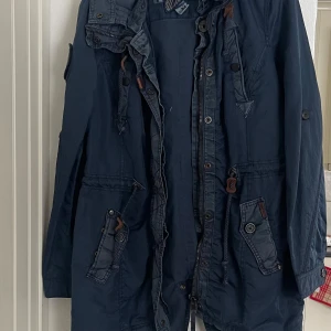 Blå parkajacka från Humör - Säljer en blå parkajacka från khujo med dragkedja och knappar. Jackan har flera praktiska fickor och justerbar huva. Perfekt för kyligare dagar med sin långa ärm och stilrena design. Köptes på zalando för 1300 använd sällan.