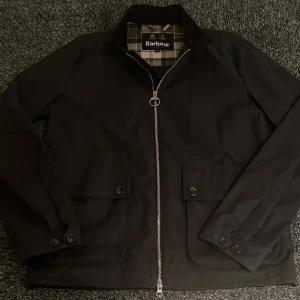 Svart jacka från Barbour - Säljer en stilren svart jacka från Barbour med klassiskt rutigt foder. Jackan har en dragkedja framtill och knappar vid ärmsluten. Perfekt för en elegant look.