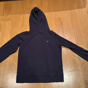 Mörkblå hoodie från GANT - Säljer en snygg mörkblå hoodie från GANT. Den har en klassisk design med en känguruficka framtill och en liten logotyp på bröstet. Perfekt för en avslappnad stil. Passar ungdomar i åldern 13-14 år. Väldigt skön och snygg. 
