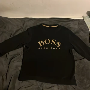 Svart tröja från Hugo Boss - Säljer en stilren svart tröja från Hugo Boss med deras logga i guld på framsidan. Tröjan har långa ärmar och en klassisk rund halsringning. Perfekt för en avslappnad men ändå elegant look.