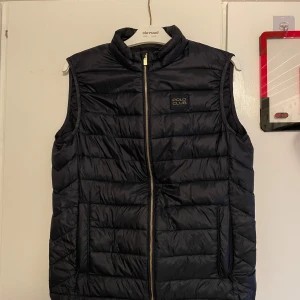 Svart dunväst från Polo Club - Snygg svart dunväst från Polo Club med dragkedja framtill. Västen har en quiltad design och är perfekt för kyligare dagar. Den har en stilren look med en diskret logga på bröstet.