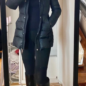 Svart quiltad dunjacka från Woolrich - Snygg svart dunjacka från Woolrich med quiltad design och pälskantad huva. Jackan har dragkedja och knappar framtill samt en badge på ärmen. Perfekt för kyliga dagar!