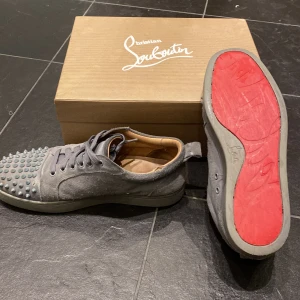 Grå sneakers med spikar från Christian Louboutin - Snygga grå sneakers i mocka från Christian Louboutin med ikoniska röda sulor. Skorna har spikar på tån och snörning framtill. Det är borta tre spikar på en sko som ni ser på bilden. Säljer eller byter!! 