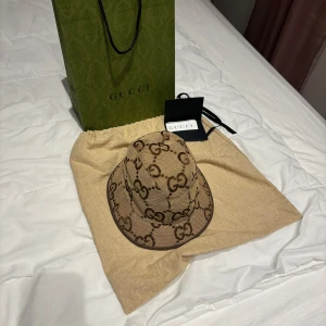 Beige mönstrad hatt från Gucci - Säljer en oanvänd, snygg beige hatt från Gucci med det ikoniska GG-mönstret. Hatten har en klassisk form och är perfekt för att ge din outfit en lyxig touch. Kommer med originalförpackning och etikett. Priset är inte hugget i sten så tveka inte på att höra av er!