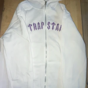 Vit hoodie från Trapstar - Säljer en vit hoodie från Trapstar med lila tryck både fram och bak. Hoodien har en dragkedja och ett coolt mönster på ryggen. Perfekt för en avslappnad stil.