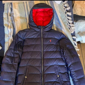 Blå och röd dunjacka från Ralph Lauren - Säljer en snygg och varm dunjacka från Ralph Lauren. Jackan är blå med röd insida och har en dragkedja framtill. Den har långa ärmar och två fickor med knappar. Perfekt för kyliga dagar! Från djur/rökfritt hem! Obs: gratis frakt!