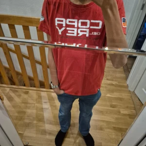 Röd Copa del Rey t-shirt - Säljer en röd t-shirt med Copa del Rey-tryck från RCNP. T-shirten har korta ärmar och flera detaljerade märken på ärmen. Perfekt för den som gillar segling och sportig stil. Den är även från sail racing 