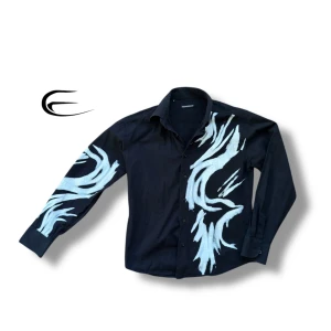 Svart skjorta med mönster från Dolce & Gabbana - Snygg svart skjorta från Dolce & Gabbana med unikt vitt mönster som sträcker sig över bröstet och ena ärmen. Skjortan har långa ärmar och knappar framtill. Perfekt för att sticka ut med stil.