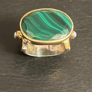 Ring med grön sten och pärldetaljer - Unik ring från Canyon med en stor oval grön sten i centrum, omgiven av guldfärgad infattning. Sidorna pryds av små pärldetaljer och ett intrikat mönster på bandet. Perfekt för att ge en elegant touch till din stil.