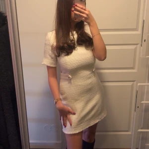 Vit kortklänning med knappar - Elegant vit kortklänning med korta ärmar och dekorativa knappar i midjan. Klänningen har en stilren design och är perfekt för en chic look. Endast använt en gång.