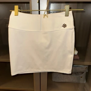 Vit kortkjol med dekorativ detalj - Snygg vit kortkjol med en liten dekorativ brosch på framsidan. Kjolen har en enkel och stilren design, perfekt för en chic look. Den är gjord i ett mjukt material som ger en bekväm passform.