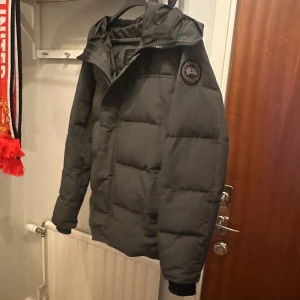 Grå dunjacka från Canada Goose med svart patch - Säljer en grå dunjacka från Canada Goose med huva och dragkedja. Jackan har en quiltad design och en svart patch på ärmen.
