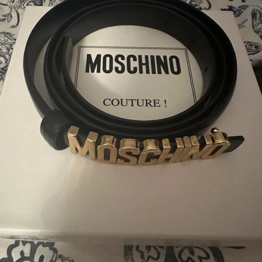Snyggt svart bälte från Moschino med ett iögonfallande guldfärgat spänne som stavar ut 'MOSCHINO'. Perfekt för att ge din outfit en lyxig touch. Bältet kommer i originalförpackning med etiketter och garantisedel. Har gjort två egan hål tillkommer bild.. Muu.