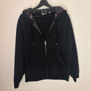 Svart hoodie från Burberry - Säljer en stilren svart hoodie från Burberry med dragkedja och justerbar huva. Den har ett diskret broderat Burberry-märke på bröstet och klassiskt rutigt mönster i huvan. Perfekt för en avslappnad look. Allt og finns kvitto påse tags. Nyskick använd 3-4 gånger endast tvättad en gång.