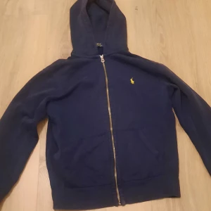Mörkblå huvtröja med dragkedja från Ralph Lauren - Säljer en snygg mörkblå huvtröja från Ralph Lauren med dragkedja och en liten gul logga på bröstet. Tröjan har långa ärmar och en praktisk huva. Perfekt för en avslappnad stil.