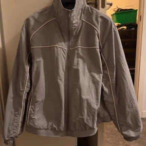 Grå vindjacka med dragkedja - Säljer en stilren grå vindjacka med dragkedja. Jackan har långa ärmar och en hög krage för extra skydd mot vinden. Perfekt för blåsiga dagar.