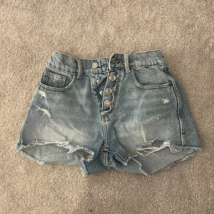 Blå jeansshorts från Zara - Säljer ett par snygga blå jeansshorts från Zara med hög midja och slitna detaljer. Shortsen har knappar framtill och klassiska femficksdesign. Perfekta för en avslappnad sommarlook!