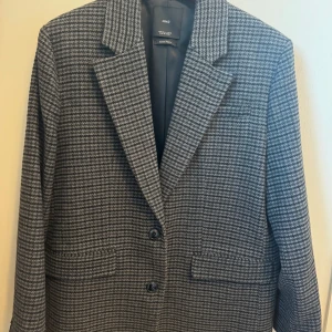 Grå rutig kavaj från Mango - Klassisk kavaj från Mango i ett grått hundtandsmönster. Kavajen har en rak passform, två knappar fram och fickor.