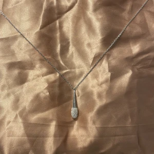 Swarovski silverhalsband - Elegant Swarovski halsband med en droppformad hängsmycke dekorerad med små stenar. Kedjan har en fin svanberlock nära låset, vilket ger en extra touch av stil.