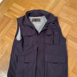 Mörkblå väst från Chevy - Säljer en mörkblå väst från Chevy med flera praktiska fickor och dragkedja. Insidan har ett rutigt mönster i grått och vitt. Perfekt för en stilren och funktionell look.