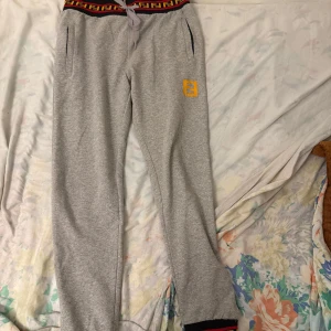 Grå mjukisbyxor med mönstrad midja - Snygga grå mjukisbyxor med en mönstrad midja i rött och svart. Byxorna har snörning i midjan och en gul broderad detalj på benet. Perfekta för en avslappnad stil.