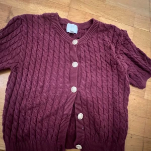 Vinröd kofta från H&M - Säljer en vinröd kofta från H&M med flätstickat mönster och dekorativa knappar. Koftan har korta ärmar och ribbade kanter. Perfekt för en mysig höstlook. 🌟