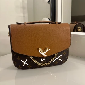 Brun och monogram handväska med kedjedetalj - Snygg handväska i brunt och monogrammönster med guldfärgad kedjedetalj. Väskan har en praktisk dragkedja och ett elegant handtag. Perfekt för att ge din outfit en lyxig touch.