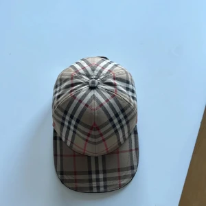 Rutig keps i beige och svart - Snygg rutig keps i beige med svarta och röda detaljer. Perfekt för en stilren look. Passar bra till både vardag och speciella tillfällen.