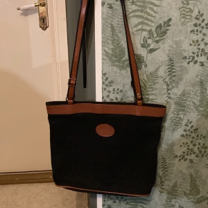 Svart väska med bruna detaljer - Svart tygväska med bruna fejkläderdetaljer. Öppnas med dragkedja och har bara ett stort fack.