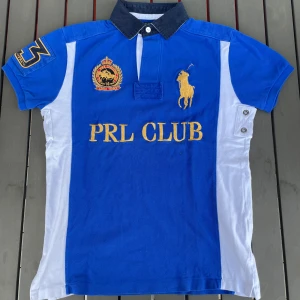 Blå och vit pikétröja från Ralph Lauren - Säljer en snygg blå och vit pikétröja från Ralph Lauren. Tröjan har korta ärmar och en klassisk krage i mörkare blått. På framsidan finns broderade detaljer med texten 'PRL CLUB' och en stor guldfärgad polospelare. Perfekt för en sportig och stilren look!