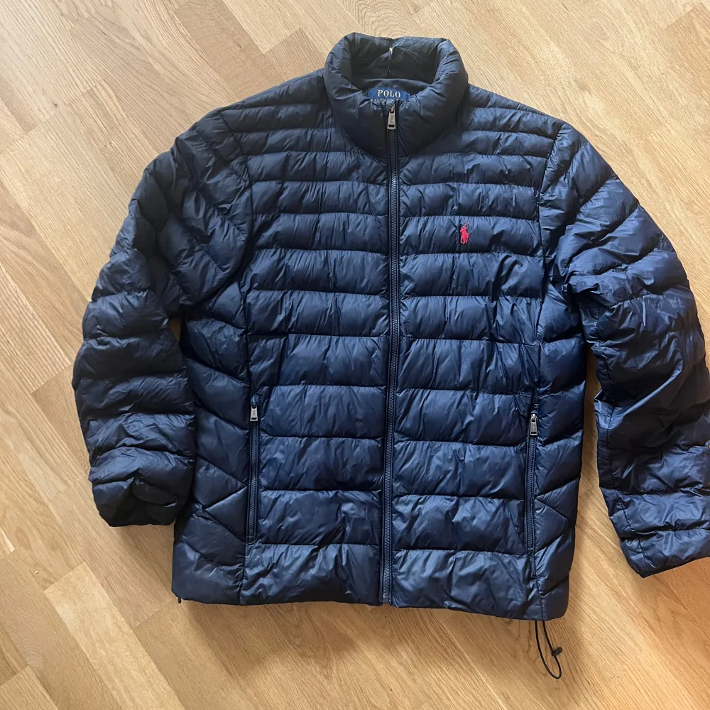 Säljer  snygg mörkblå pufferjacka från Polo Ralph Lauren. Jackan har en broderad röd logga på bröstet och är perfekt för höst och vinter. Den är långärmad och har en dragkedja framtill. Jackan är i nyskick. Jackan är i herr strl. Nypris 4300:-. Jackor.