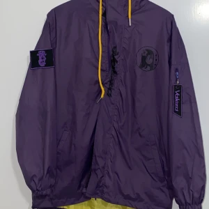 Lila vindjacka med tryck - Säljer en cool lila vindjacka från Volezca med unika tryck. Jackan har en stor grafisk design på ryggen med texten 'Missing Pieces' och en illustration av en tjej med en gul mask. På framsidan finns en mindre tryck av en tjej med en blomma i håret. Jackan har en huva med gula dragsnören och elastiska ärmslut. Perfekt för dig som vill sticka ut!