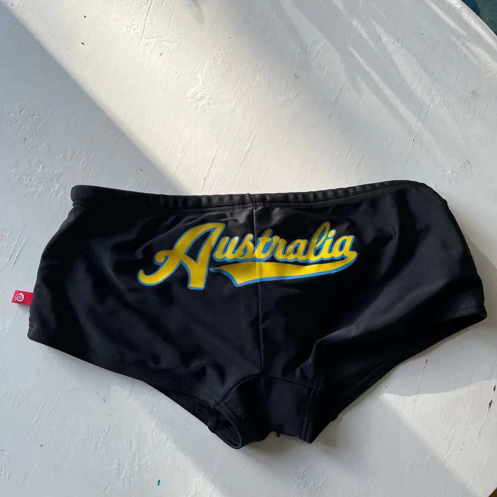 Säljer ett par snygga svarta badshorts från AussieBum med texten 'Australia' i gult och blått på framsidan. De är gjorda av 80% nylon och 20% spandex, vilket gör dem superbekväma och perfekta för en dag på stranden. Shortsen har en tight passform och en liten röd etikett på sidan. Perfekta för sommarens alla badäventyr! 🏖️. Shorts.