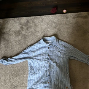 Blå och vit randig skjorta från Zara - Säljer en snygg blå och vit randig skjorta från Zara i storlek 11-12 år. Skjortan har långa ärmar och knappar framtill. Perfekt för både vardag och festliga tillfällen. Den är i bra skick och redo för en ny ägare!