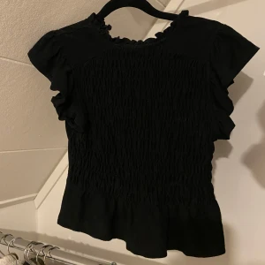 Svart topp från Miss Selfridge - Säljer en svart topp från Miss Selfridge med korta ärmar och volangdetaljer. Blusen har en smockad design som ger en fin passform och en liten volangkant vid halsen🩷